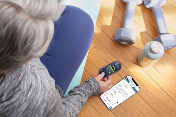 Ältere Frau beim Sport hält ein OneTouch Blutzuckermessgerät in der Hand, neben ihr ein Smartphone mit der OneTouch Reveal App, Hanteln und eine Trinkflasche auf dem Boden